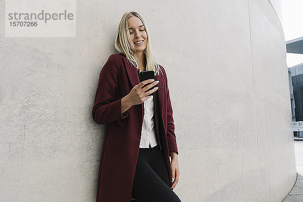 Blonde Geschäftsfrau  die ein Smartphone benutzt und sich an eine Wand lehnt