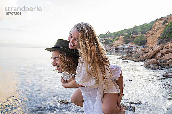 Junger Mann nimmt seine Freundin huckepack vor dem Meer mit  Ibiza  Balearen  Spanien