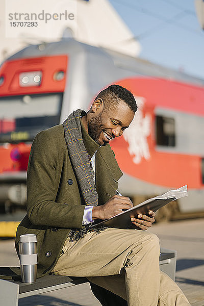 Porträt eines stilvollen Geschäftsmannes mit wiederverwendbarer Tasse und Dokumenten  die auf den Zug warten