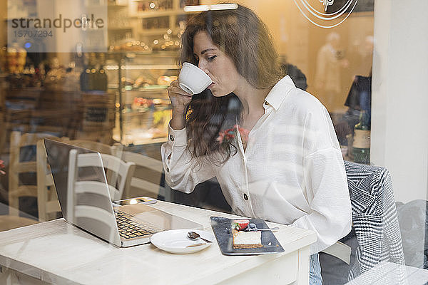 Frau mit Laptop trinkt Kaffee hinter einer Fensterscheibe in einem Cafe