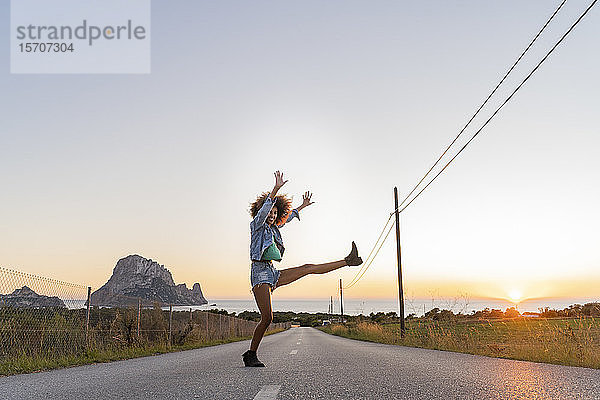 Junge Frau  die bei Sonnenuntergang auf der Straße steht und ihr Bein hebt  Ibiza