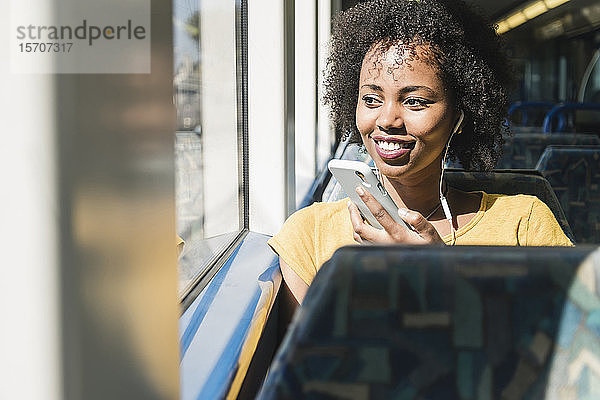 Lächelnde junge Frau mit Kopfhörern und Smartphone in einem Zug
