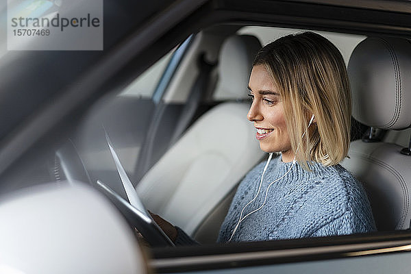 Lächelnde junge Frau mit Dokumenten  Tablett und Kopfhörern in einem Auto
