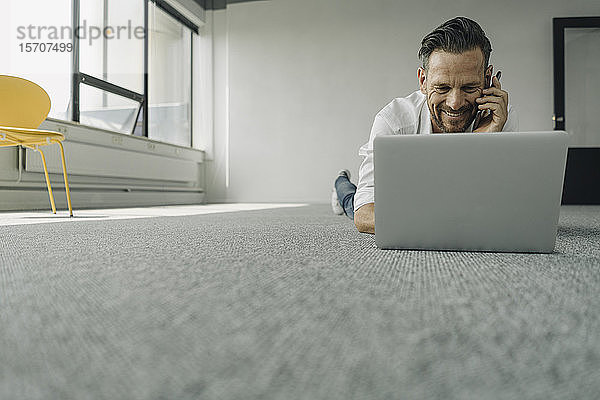 Lächelnder reifer Geschäftsmann  der in einem leeren Büro mit Laptop und Smartphone auf dem Boden liegt