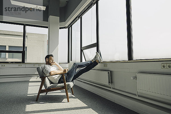 Reifer Geschäftsmann mit Laptop entspannt sich am Fenster im leeren Büro