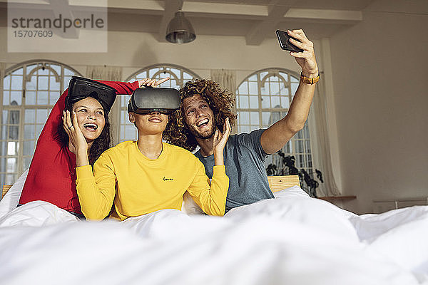 Glückliche Freunde  die zu Hause im Bett mit VR-Brille und Smartphone Spaß haben
