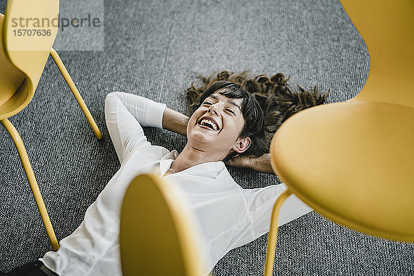 Lachende Geschäftsfrau  die in einem Büro auf dem Boden zwischen Stühlen liegt