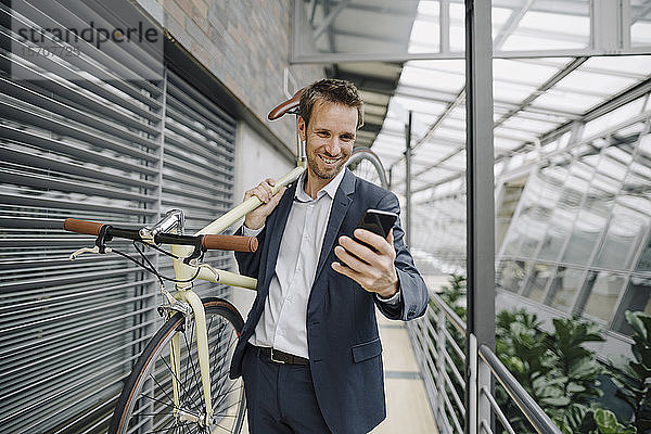 Lächelnder Geschäftsmann mit Handy mit Fahrrad in modernem Bürogebäude