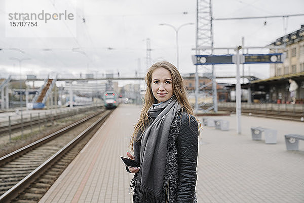 Porträt einer lächelnden jungen Frau mit Smartphone auf dem Bahnsteig stehend