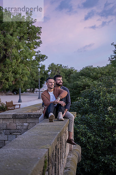 Lächelndes schwules Paar sitzt bei Sonnenuntergang an einer Wand