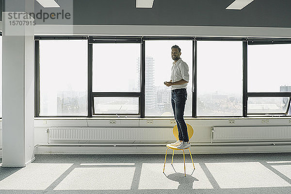 Glücklicher reifer Geschäftsmann steht auf gelbem Stuhl in leerem Büro