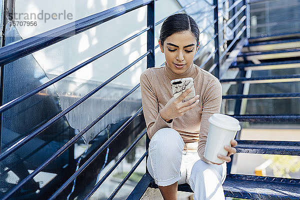 Junge Frau mit Getränk zum Mitnehmen  die auf einer Außentreppe sitzt und ein Smartphone benutzt
