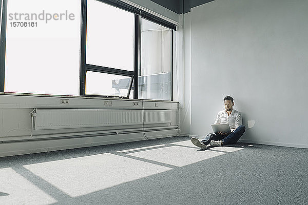 Ein reifer Geschäftsmann sitzt in einem leeren Büro auf dem Boden und benutzt einen Laptop