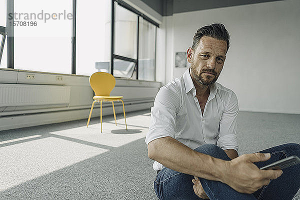 Porträt eines reifen Geschäftsmannes  der in einem leeren Büro auf dem Boden sitzt und ein Handy in der Hand hält