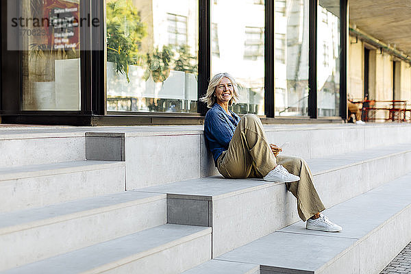 Reife Geschäftsfrau  die auf Stufen in der Stadt sitzt und ein Smartphone benutzt