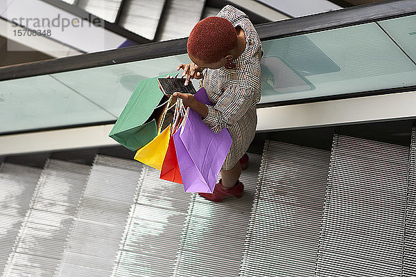Junge Frau hält bunte Einkaufstaschen in der Hand und überprüft ihr Handy beim Treppabgehen