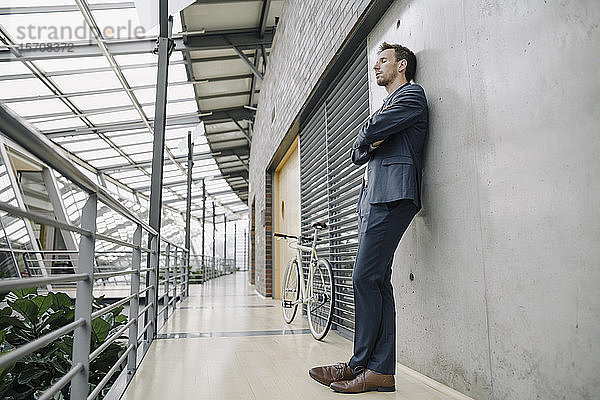 Geschäftsmann mit geschlossenen Augen an eine Wand gelehnt in einem modernen Bürogebäude