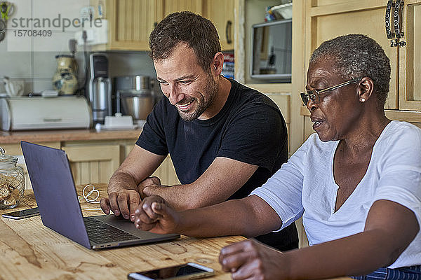Ältere Frau und lächelnder Mann sitzen am Küchentisch und teilen sich einen Laptop