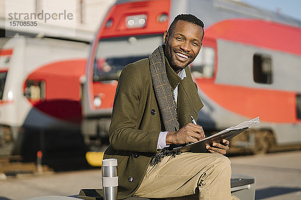 Porträt eines glücklichen Geschäftsmannes mit wiederverwendbarem Becher und Dokumenten  der auf den Zug wartet