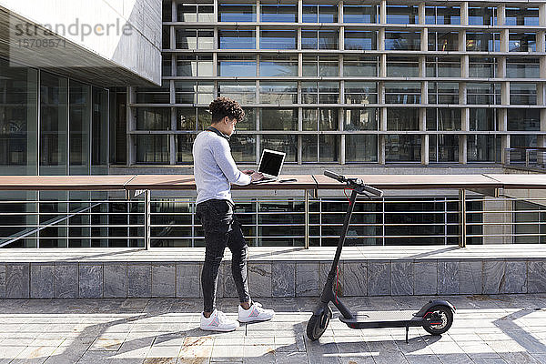 Junger Mann mit E-Scooter mit Laptop in der Stadt