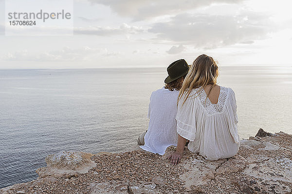 Rückenansicht eines jungen Paares  das auf einem Felsen vor dem Meer sitzt  Ibiza  Balearen  Spanien