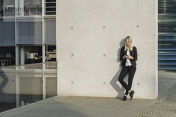 Blonde Geschäftsfrau mit Smartphone an der Wand gelehnt