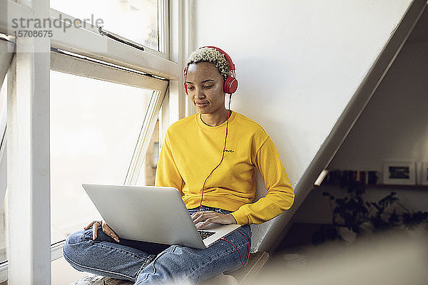 Junge Frau sitzt zu Hause am Fenster mit Laptop und Kopfhörern