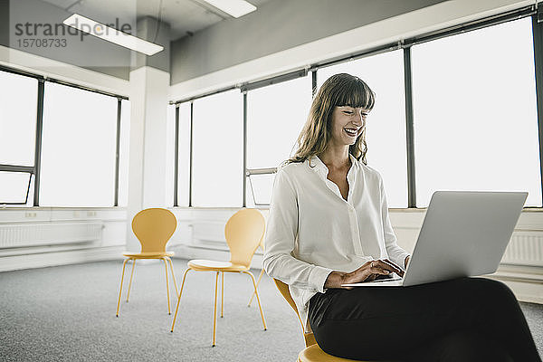 Lächelnde Geschäftsfrau sitzt mit einem Laptop auf einem Stuhl in einem leeren Büro