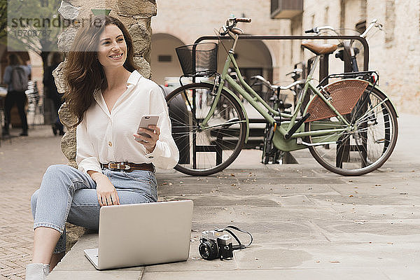 Lächelnde junge Frau mit Smartphone  Kamera und Laptop im Freien