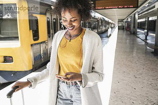Lächelnde junge Frau mit Kopfhörern und Smartphone am Bahnsteig