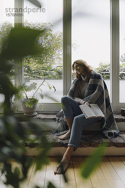 Eine reife Frau sitzt auf einer wunderbaren Fensterbank  in eine Decke gewickelt  liest ein Buch