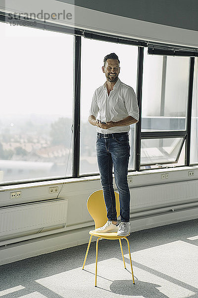 Glücklicher reifer Geschäftsmann steht auf gelbem Stuhl in leerem Büro
