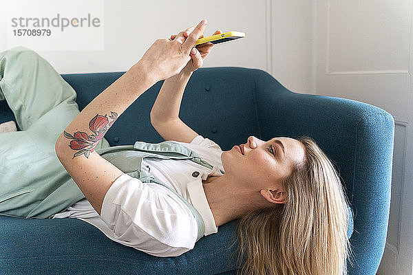 Junge Frau liegt auf Couch und benutzt Mobiltelefon
