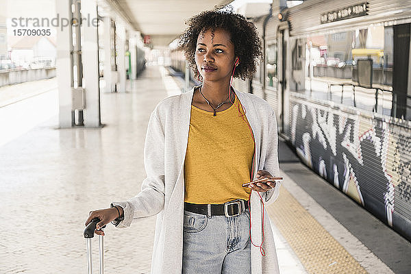 Junge Frau mit Kopfhörern und Smartphone am Bahnsteig schaut sich um