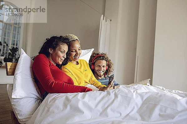 Glückliche Freunde entspannen zu Hause im Bett mit Smartphone