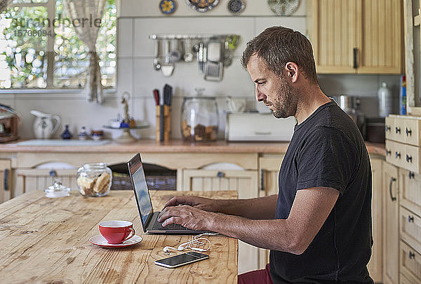 Mann arbeitet von zu Hause aus  sitzt am Küchentisch  benutzt Laptop und Smartphone