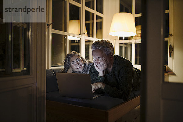 Glückliches älteres Paar  das abends zu Hause auf der Couch liegt und einen Laptop benutzt