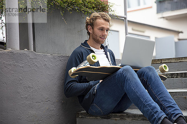 Junger Mann sitzt auf Stufen und benutzt Laptop auf Longboard