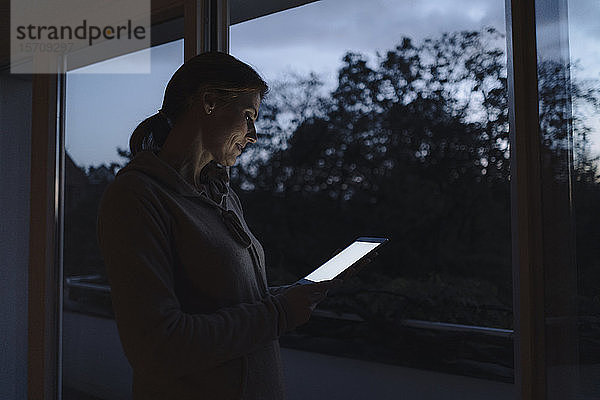 Frau mit digitalem Tablett  in der Dämmerung am Fenster stehend