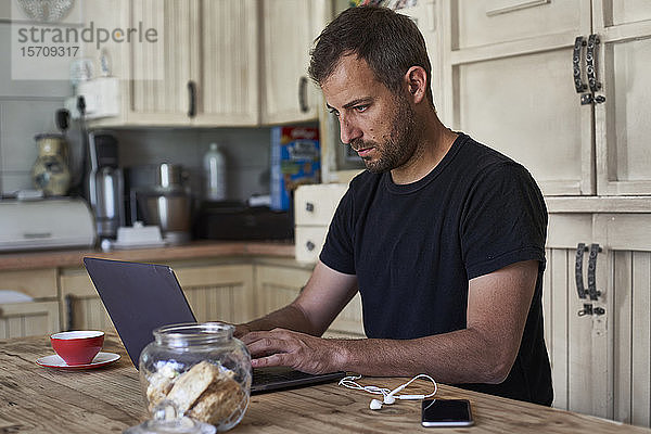 Mann arbeitet von zu Hause aus  sitzt am Küchentisch  benutzt Laptop und Smartphone