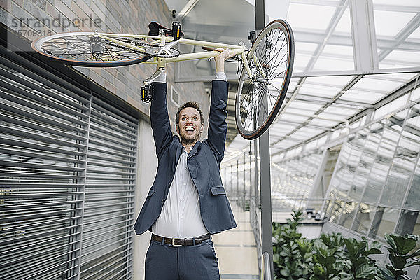 Fröhlicher Geschäftsmann hebt Fahrrad in modernem Bürogebäude hoch