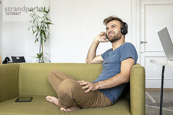 Lächelnder Mann sitzt im Büro auf der Couch und hört Musik