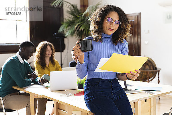 Frau mit Papier und Kaffeetasse im Büro mit Kollegen im Hintergrund