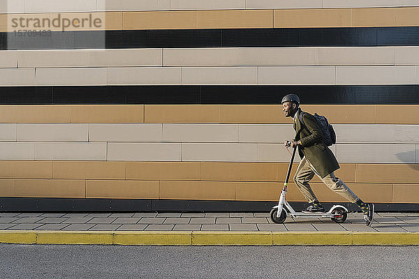 Stilvoller Mann mit Helm und Roller fährt an einem Gebäude vorbei