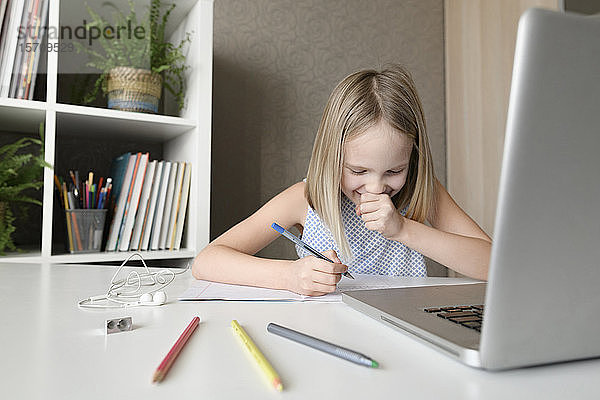 Lachendes Mädchen sitzt zu Hause am Tisch  macht Hausaufgaben und benutzt den Laptop
