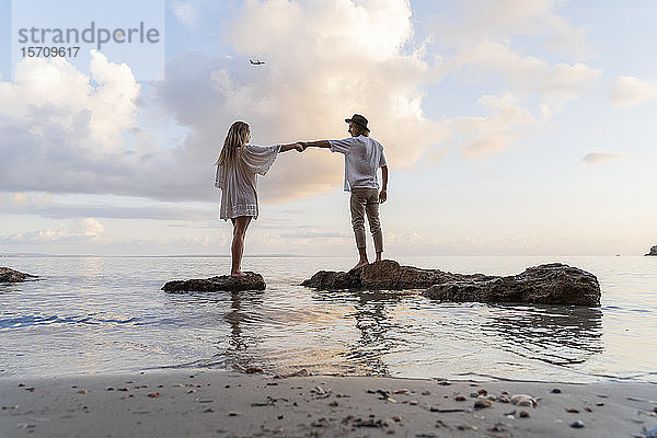 Junges Paar steht auf Felsen vor dem Meer und hält sich an den Händen  Ibiza  Balearen  Spanien