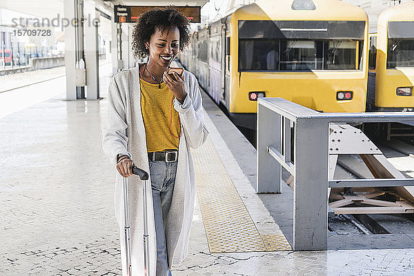 Lächelnde junge Frau mit Kopfhörern  die ein Smartphone am Bahnsteig benutzt