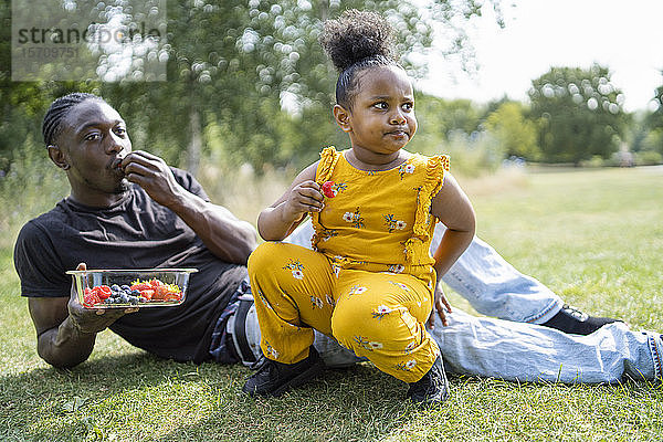 Vater und Tochter essen Obst auf einer Wiese in einem Park