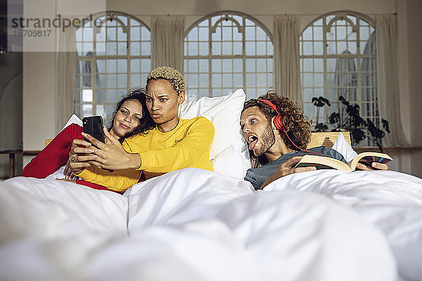Glückliche Freunde haben zu Hause mit Smartphone Spaß im Bett
