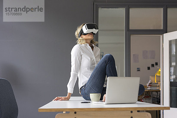 Junge Geschäftsfrau sitzt im Büro auf dem Schreibtisch und trägt eine VR-Brille